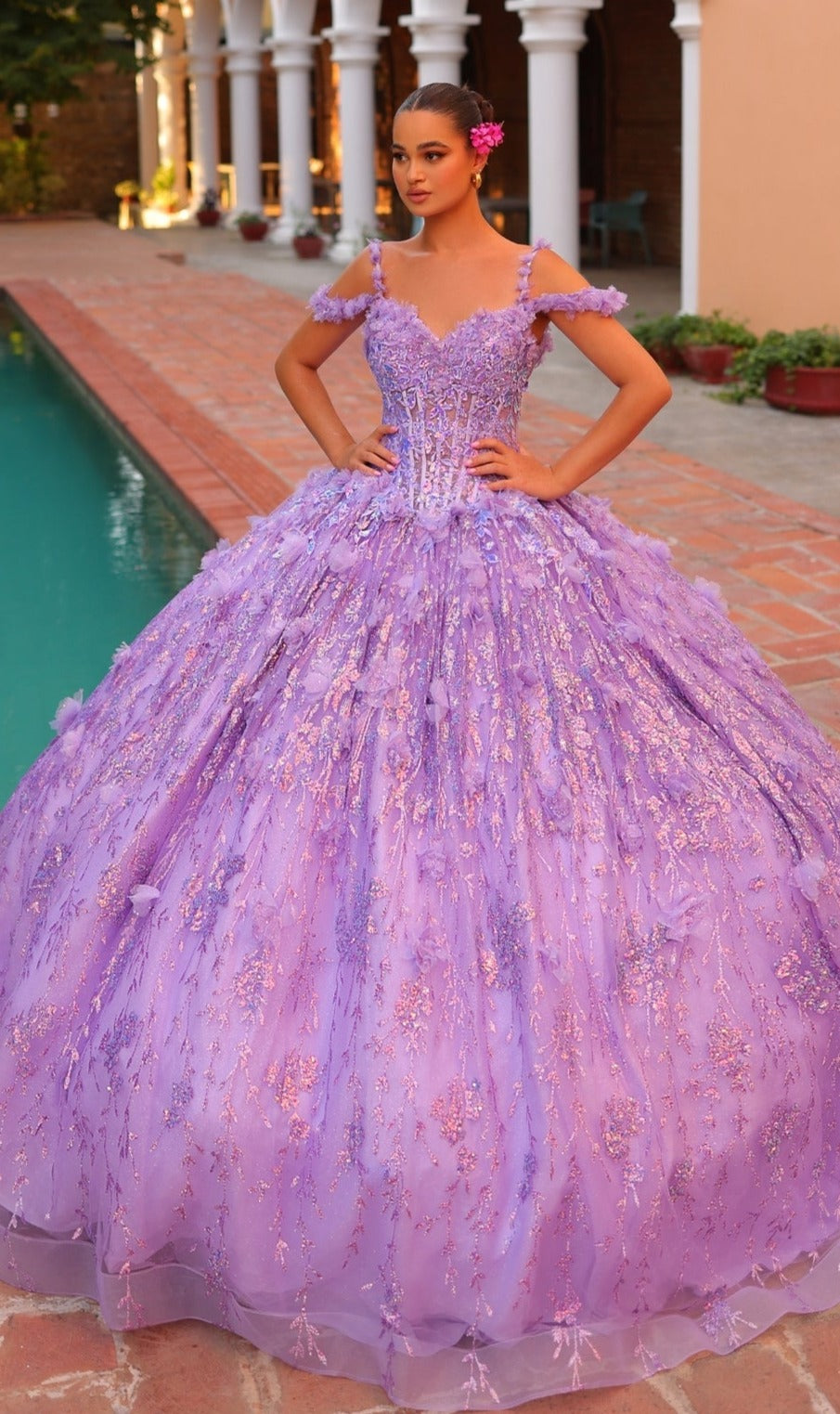A young woman poses by a pool in the Quinceanera Dress 54320 By Amarra, featuring voluminous lavender layers, off-the-shoulder straps, and 3D floral details. She stands confidently with her hands on her hips and a pink flower in her hair.